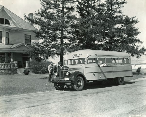 International D-30 bus with 19-foot Hicks body picking up two young men for school. The young (teenaged) men are wearing light jackets and brimmed hats. The side of the bus reads: "Monroe County School."