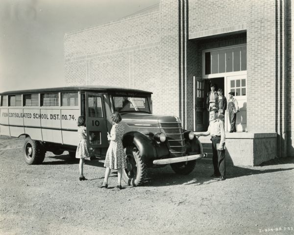 Students boarding an International D-35 bus with a Picayune body style and 179-inch wheel base. The bus belonged to the Fox Consolidated School District, No. 74 and is parked in front of what appears to be a school building.