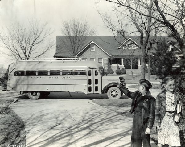 Two children walking up the driveway while waving back at a school bus sitting at the curb in a residential neighborhood. The bus is a 22 foot International D-35 school bus with a Hicks body. The side of the bus reads: "Rockland School District."
