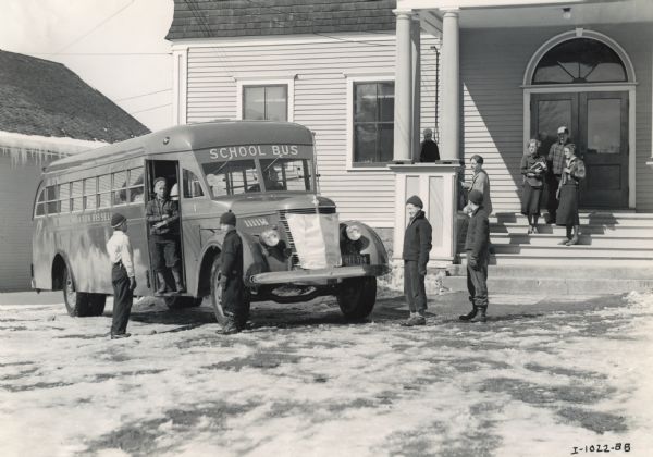 Photograph of students disembarking an International D-40 school bus at Newcomb Central School. The students appear to be dressed for winter: coats, hats, mittens. This bus is equipped with a Rex-Watson body, 215-inch wheel base, and seats 49 passengers. The side of the bus is labeled: "L. Morton Bissell".