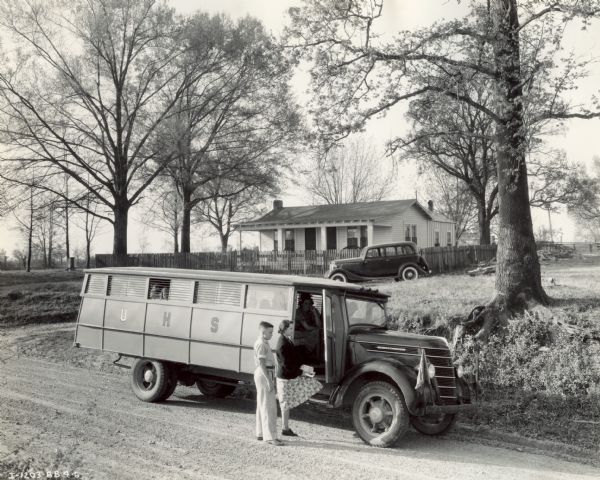 International D-30 bus with Picayune body picking up students for school. There is an automobile in the yard in the background. This bus was equipped with an 18 foot Picayune body and 173-inch wheel base.