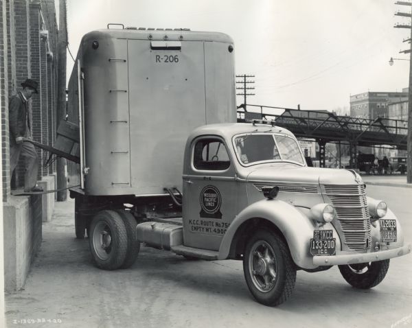 View along side of a building of a man unloading/loading boxes from a loading dock. An International DS-30 truck-tractor with semi-trailer owned by Columbia Terminals Company is parked alongside the building with its side door opened. The truck was operated by the Missouri Pacific railroad service.