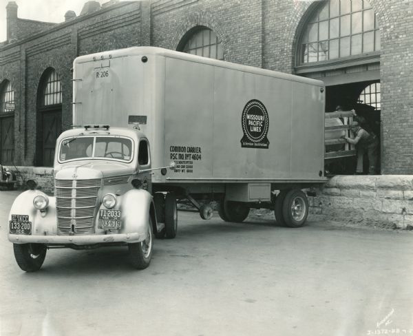 Three-quarter view of an International DS-30 truck-tractor with semi-trailer owned by Columbia Terminals Company. The truck was operated by the Missouri Pacific railroad service. Men on a loading dock are loading/unloading an object packed in a large open crate.