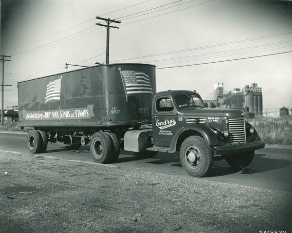 International KR-11 truck on a paved road. The trailer is decorated with two large images of the U.S. flag. Original caption reads: "FREIGHT HAULING International Model KR-11, 161-inch wheel base. Axel Ratio- 7.10 to 1. Transmission- F-54, overdrive in fifth. Semi-Trailer. Payload- 18 to 22 tons. Distance- varies with trip. Fuel Consumption- 6 miles per gallon. Owner- Endres Delivery, Inc., Buffalo, N.Y. (owns 35 Internationals)." Side of truck reads: "Hasten Victory--BUY WAR BONDS and STAMPS."