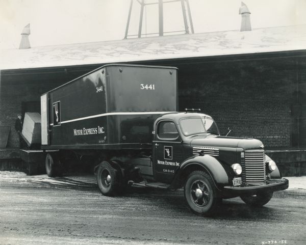 Man loading a box into an International K-7 truck with semi-trailer, operated by Motor Express, Inc. The truck is backed up to the loading dock of a building.
