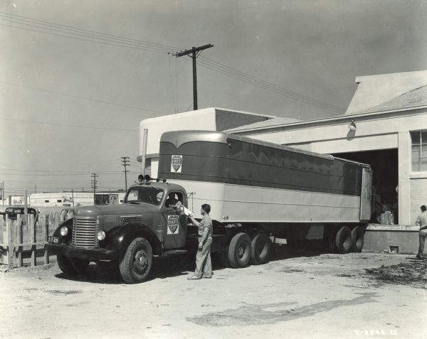 International KRD-11 truck with a 35 foot Trailmobile closed-body semi-trailer (back 10 ft. removable for loading with crane); 161 inch wheelbase; and Brown-line transmission. The truck was operated by Charles P. Hart Transportation Co. A man is standing next to the truck talking with the driver.