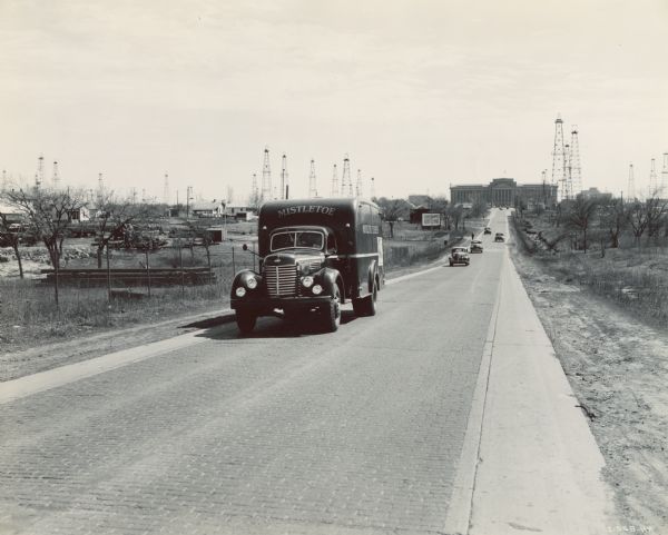 International highway truck operated by Mistletoe Express Service, Inc. Original caption reads: "The truck shown was traveling on a busy highway leading out of Oklahoma City. The state house is shown in the background with oil derricks everywhere, even on the capitol grounds and on the grounds of the governor's mansion nearby. Green above and black below the belt line are the prevailing colors with gold lettering and trim."