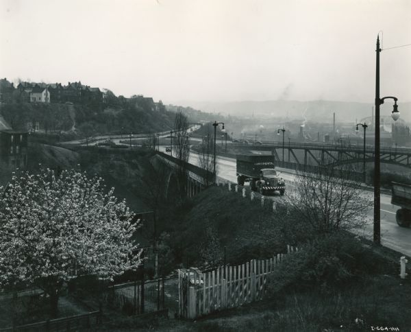 Continental Transportation Lines, Inc. operating an International semi-truck on a highway with an industrial area in the background. Original caption reads: "Because they are hauling commodities which are 90 percent directed to the war effort, Continental was recently successful in obtaining releases from the Office of Defense Transportation and War Production Board at Washington D.C., for the purchase of 10 International Model KS and KR-11 truck tractors. One of these is shown in accompanying illustrations on the Ohio River Boulevard and McKees Rock Bridge with industrial Pittsburgh in the background . . . These International tractors are hauling 28-foot van-type trailers with capacity loads authorized by various states. Both are painted a rich color red."