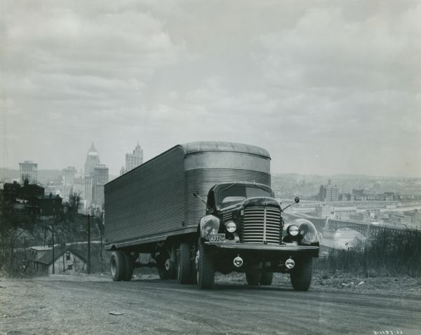 View from side of a road of an International KR-12 truck equipped with 149 inch wheel base and semi-trailer. There is a cityscape in the background.