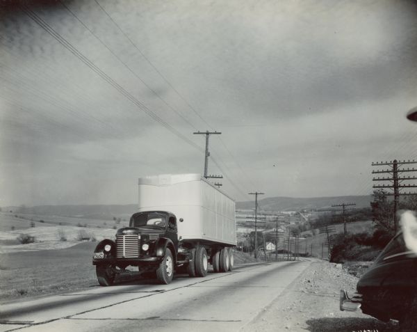 View from the side of a road of an International KR-12 truck equipped with 149 inch wheel base and semi-trailer. In the foreground on the right is a parked automobile. In the background are fields, and power lines run along the road.
