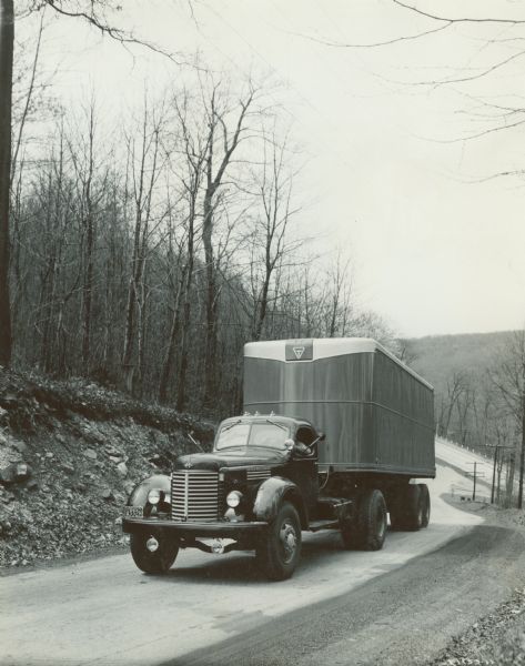View from the side of a road of an International KR-12 truck equipped with a 149 inch wheel base and semi-trailer. The road is in a wooded, hilly region.