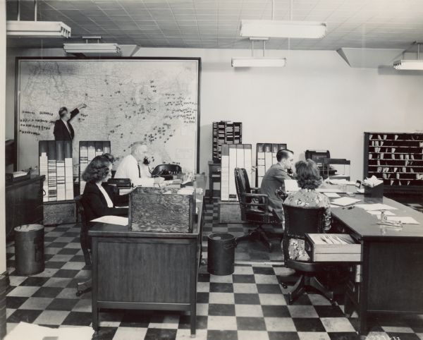 Traffic control room of Aero Mayflower Transit Company of Indianapolis. Men and women are seated at desks in an office setting. A few of the employees are using telephones. One man stands at the back of the room plotting something on a large United States map.