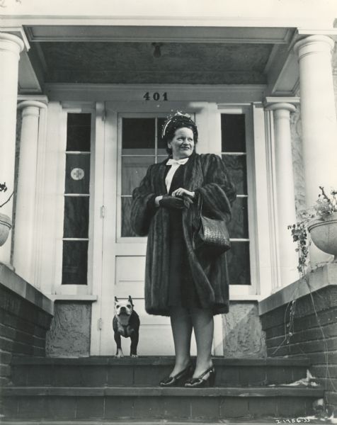 A woman is standing on the front steps of a house with a french bulldog. The woman is dressed up in a fur coat, a fancy hat and high heels.