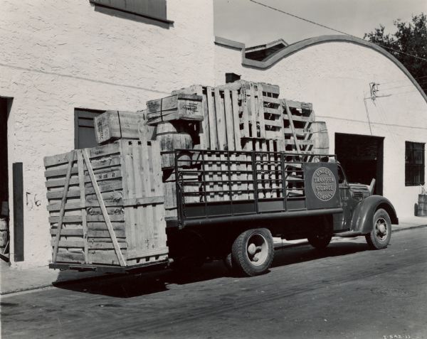 Three-quarter rear view of an International K-6 truck loaded with wooden crates and barrels. Original caption: "This latest purchase, a K-6 International, made by the Union Transfer & Storage Company of Miami, Florida, is part of a fleet of seven units, four of which are Internationals. The truck shown is equipped with all-metal, 14-foot stake body with an Anthony hydraulically operated tail lift. [This photograph] shows the truck with tail and load on it lowered. Length of body including this tail life is 17 feet."