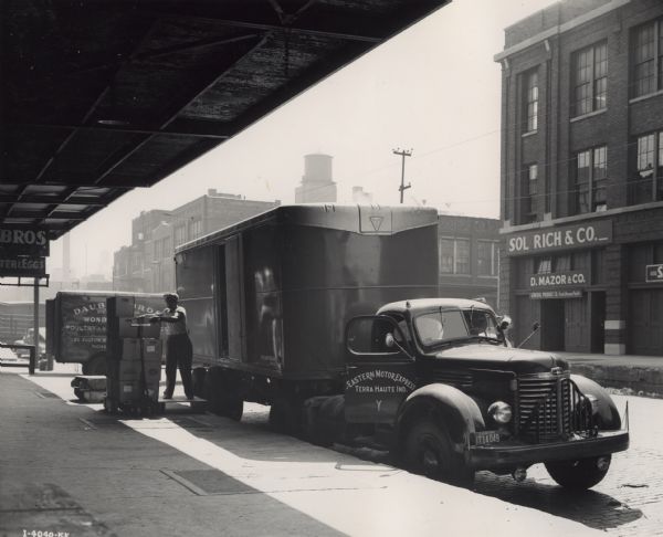 A man unloads boxes from an International KB-10 truck parked on a city street. The sidewalk appears to be under an elevated railroad track or some time of awning. The original caption reads: "An International KB010 leased from the Eastern Motor Express to Elden Strange."