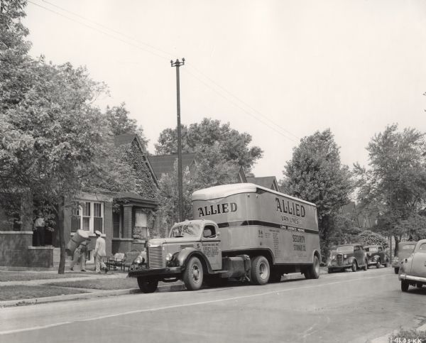 View from across a residential street of a moving scene. Men are loading household items into an International KB-47 truck owned by Security Storage Company of Duluth, Minnesota. The truck was equipped with a Fruehauf body. The side of the van reads: "Allied Van Lines, Inc. Long Distance Moving" and "Security Storage Co."