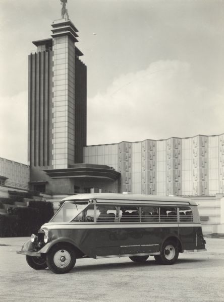 Man in driver's seat of an International bus parked in front of a building with a tower.