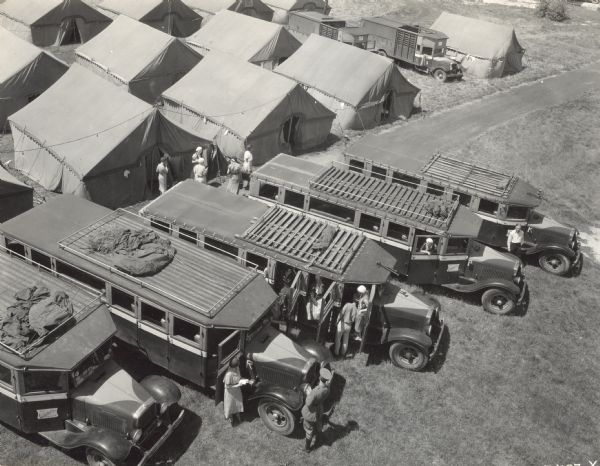 View from above of five International buses labeled, "The Omnibus College" parked in front of a group of tents. There appear to be men in uniform standing near the buses, as well as women who are disembarking from the buses.