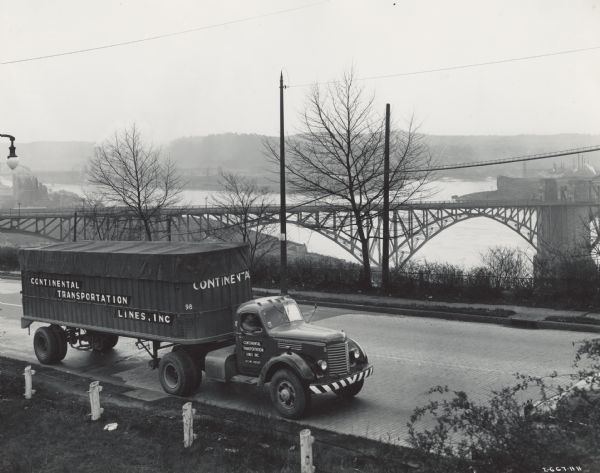Continental Transportation Lines, Inc. operating an International semi-truck (tractor-trailer) on a road with a hazy view of a bridge, a body of water and some industrial buildings in the background. Original caption reads: "Because they are hauling commodities which are 90 percent directed to the war effort, Continental was recently successful in obtaining releases from the Office of Defense Transportation and War Production Board at Washington D.C., for the purchase of 10 International Model KS and KR-11 truck tractors. One of these is shown in accompanying illustrations on the Ohio River Boulevard and McKees Rock Bridge with industrial Pittsburgh in the background and also at the Pittsburgh terminal. These International tractors are hauling 28-foot van-type trailers with capacity loads authorized by various states. Both are painted a rich color red."