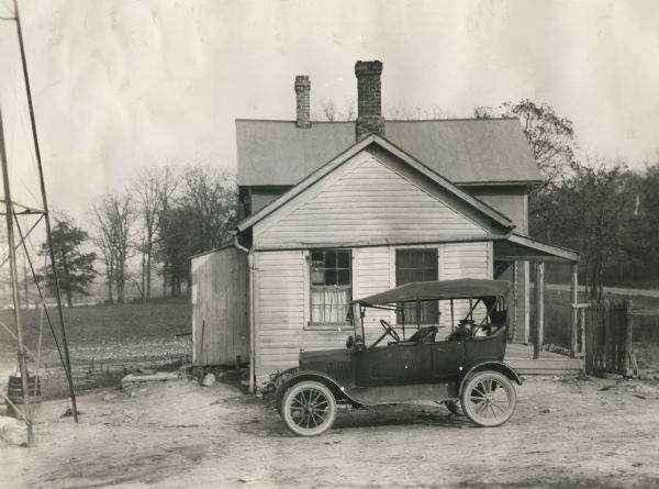 Automobile parked outside a rural house. Milk cans are stowed in the back of the automobile. The portion of a base of what is probably a windmill is on the left.