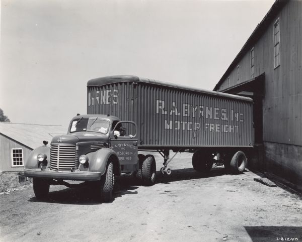 Man in driver's seat with the door open backing an International KR-11 with semi-trailer up to a loading dock at Hurff Cannery. The truck was operated by R.A. Byrnes Motor Freight Company. According to the original caption: the truck was painted red.