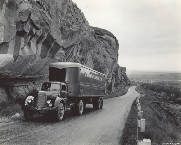 International KR-11 truck with semi-trailer (semi-truck) operated by Northwest Freight Lines. The truck is on a mountain road next to a large rock formation. Original caption reads: "A recent purchase obtained through ODT release is the KR-11 International motor truck shown traveling up scenic Rim Rock Highway with Billings [Montana] in the background... The Refer-van trailer is 32 feet long, 8 feet wide and 7 feet high.