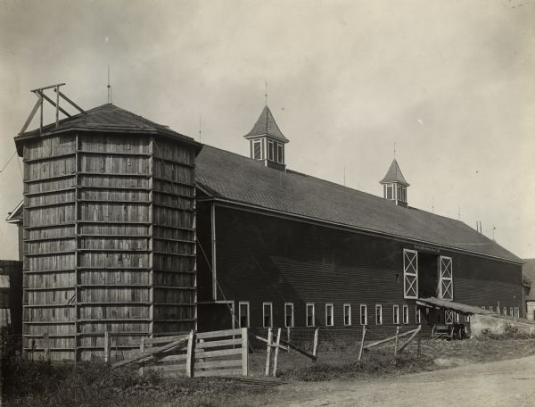 Dairy barn owned by Schneider Bro's. dairy with a large ramp leading up to the open barn door. There are two gable vents on the roof of the barn. A wagon is parked below the ramp. There are various styles of fencing around the barn. On the left end of the barn is a wooden silo.