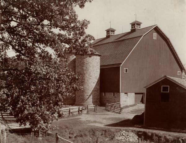 Dairy barn and glazed hollow tile silos on M.A. Browning's dairy farm. There are two gable vents on the roof of the barn. There are multiple types of fencing on the farm and other farm buildings.