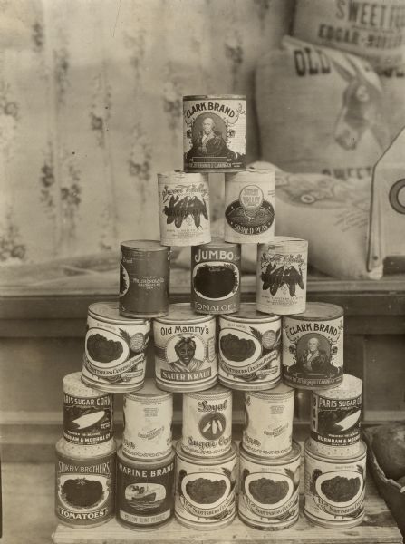 A pyramid of stacked canned goods at an Alabama store. Original caption reads: "The people of Alabama are artistically inclined as evidenced by the car loads of fancy labeled canned goods packed in other states and shipped to them for retail at high prices. In this sidewalk pyramid which was photographed in front of J.E. Hasson & Company store, Aliceville, Feb. 15, 1915, were cans of tomatoes from Newport, Tenn., which retail at fifteen cents per can and some from Baltimore which peddle at ten cents per can. Sour kraut from Scottsburg, Indiana, brings fifteen cents per can and small cans at that. Sugar corn from Gibson City, Illinois, and from Portland, Maine, retail alike for ten cents per can, while soaked peas from Baltimore are the same price. Peaches from Winters, California, bring fifteen cents per can, but pie peaches from Baltimore could be obtained from this exhibit at ten cents per can. Lye  hominy from Jeffersonville, Indiana, were being bought in large quantities by Aliceville residents at fifteen cents per can, and it was worth the price for each can has a highly colored lithograph of George Rogers Clark set in scroll, as shown on top can. Pet cream from Highland, Ill., also was a fine seller at this store and sold for ten cents a can straight. Evidently all dogs around Aliceville must have died if conditions were similar to one farm visited by some of the speakers who found canned milk on the table. Considerable surprise was expressed by the visitors to which the hostess replied, 'We haven't had any fresh milk since the dog died.' Pressed for an explanation she said that the dog used to bring up the cow morning and night to be milked, but since the dog died, the cow had not come up and no one at the house wanted to run their legs off trying to drive her up, so they were buying canned milk."