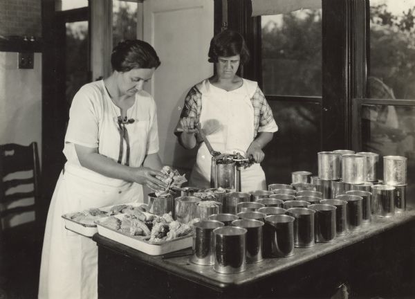 Two women working at P.G. Holden's Farm canning chicken. On the table there is canning equipment, chicken, and open cans.