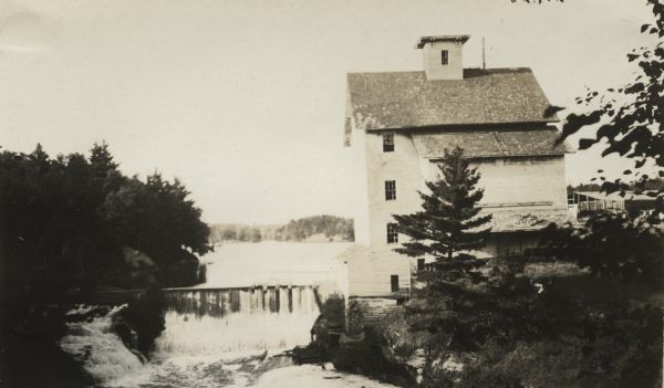 View across river of an "old mill." There is a waterwheel attached to the left side of the mill. A small waterfall and a number of trees and foliage surround the area.