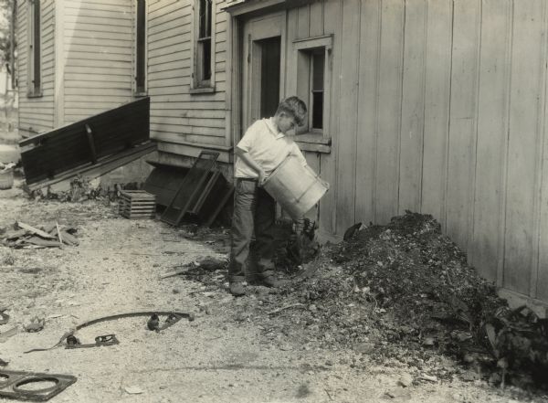 A boy dumping ashes against the side of a house. This photo was taken for International Harvester's Agricultural Extension Department to illustrate potential farm hazards.