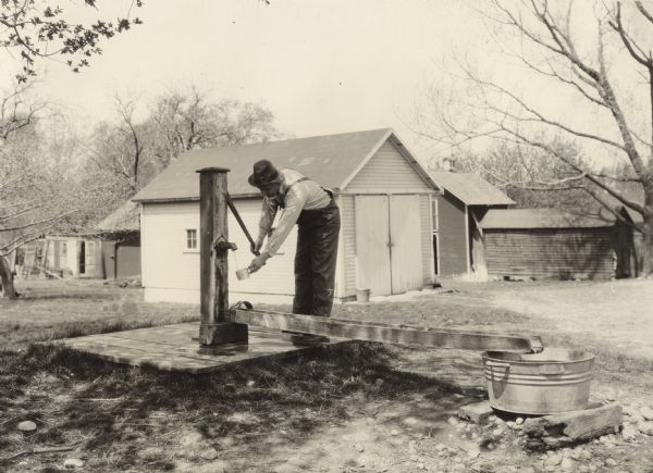 A man pumps water from an old wooden pump into a tin cup. This photo was taken for International Harvester's Agricultural Extension Department to illustrate potential farm hazards.