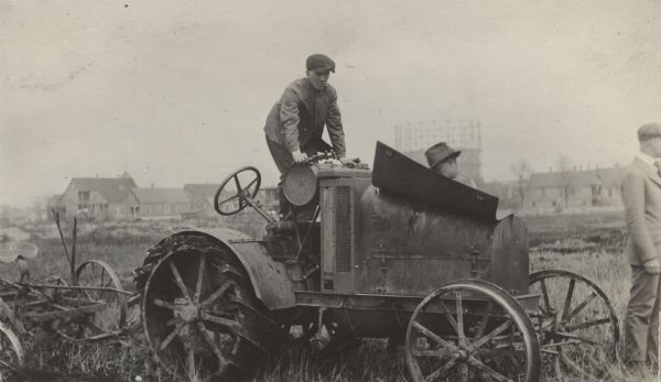 Three men in a field working on an International 8-16 tractor. One of the men is dressed in a suit. There are houses in the background.
