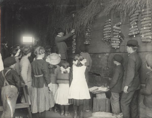 A man is hanging up strings of corn to demonstrate corn testing in front of a group of children (students?).