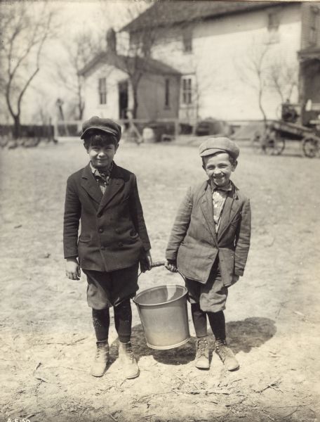 Two boys holding a pail of water. There is a wagon parked near a building in the background. Original caption reads: "Bringing a pail of warm water for the rag doll tester."