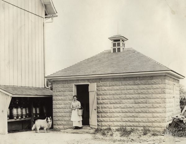 A woman and her dog standing at the doorway of a concrete block milk house.