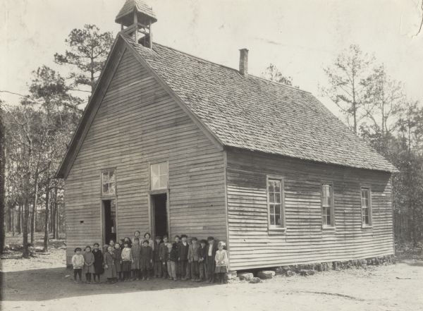 Small schoolhouse with a bell tower on the roof over the entrance to the building. The schoolhouse is surrounded by trees. A class of students is pictured posing in front of the school. Original caption reads: "Providence School near Vinemont, Alabama... 130 children up to 16-17 enrolled. No toilets. Teacher says they have always gotten along just going into the woods in back. Situation said to spread hookworm disease."