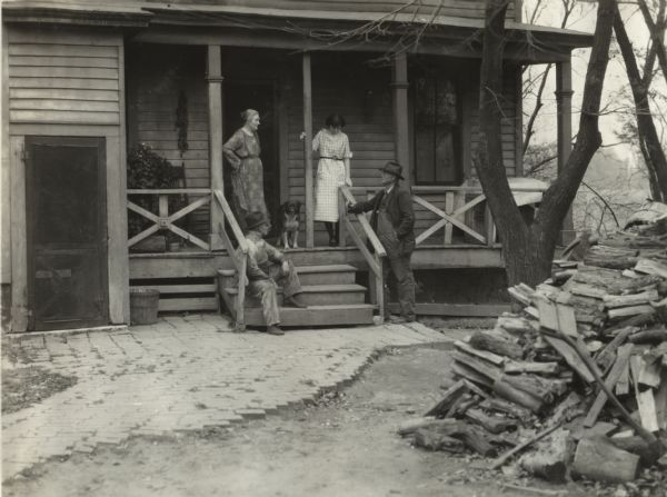The back porch and the nearby wood pile, on the John Esch farm. There are two men, two women and a dog on the porch.