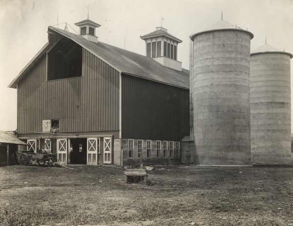 Dairy barn with two silos on the right side. There is a wagon in the front of the barn and a ladder which leads up into the second floor. Hay is visible in the open doorway of the barn. Original caption reads: "Dunham's dairy farm near Wayne, Ill."