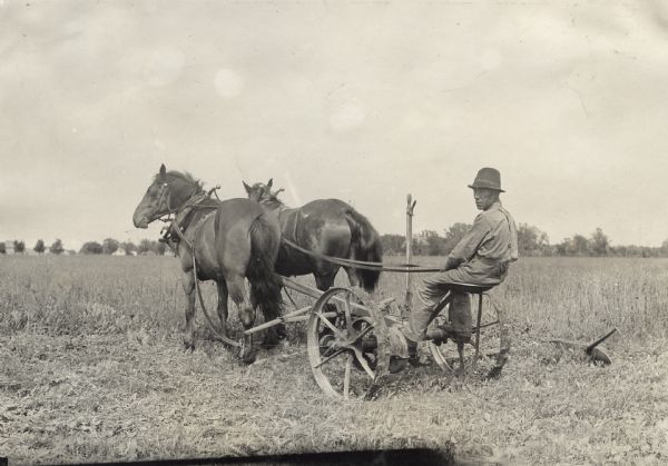 Man operating a horse-drawn mower in a field. The horse on the left is wearing blinders.