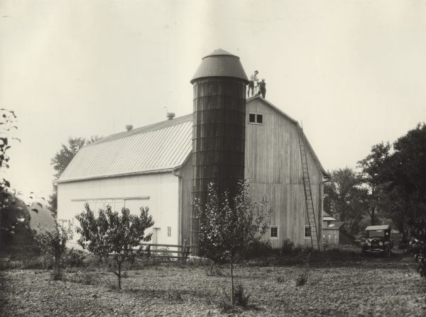 Two men are on top of a barn at an orchard, taking a panoramic photograph of an orchard.