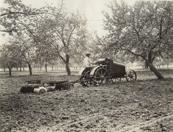 Man pulling a disc harrow in an orchard with an International 8-16 tractor. The original caption reads, "used in 'Battle the Bugs'."
