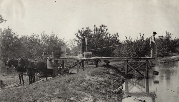Three men in an orchard mixing up chemicals to spray the trees. One of the men is on the back of a horse-drawn wagon with a barrel. The other two men are standing on a pier: one appears to be mixing something in a large tub, the other is fetching water with a pail from the end of the pier.
