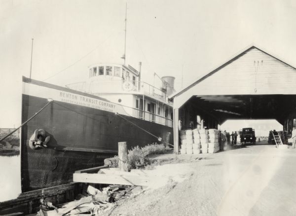 Large cargo ship operated by the Benton Transit Company. On the dock near an open shed are many bushels of fruit, including melons and tomatoes (according to the caption: "Fruit: melons and tomatoes").