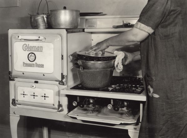 Woman steaming spinach in a hot-water bath on a Coleman Pressure Range at the Harvester Farm. The image is part of a series illustrating canning methods.