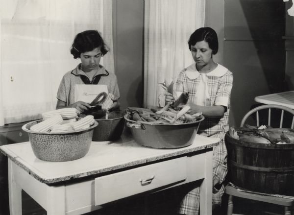 A woman and a girl sitting at a table, husking and silking corn at the Harvester Farm. The image is one of a series illustrating canning methods.