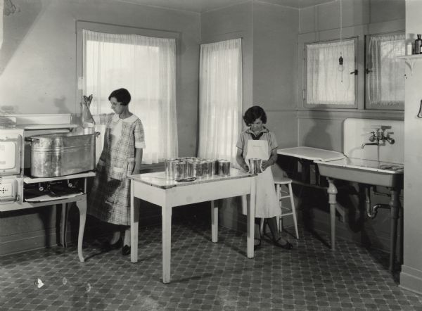 A woman and a girl standing in a kitchen. They are in the process of canning corn at the Harvester Farm, and are removing cans from the cooker and allowing them to cool.