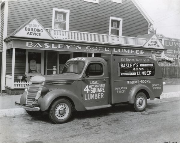 An International D-2 truck owned by Basley's Good Lumber Company parked in front of the company storefront. The text on the truck reads, "Use Weyerhaeuser 4-Square Lumber," "Call Newton North 5500," "Basley's Good Lumber. Roy S. Edwards. You Can Depend On Us," "Windows-Doors, Insulation, Fences," and "29 Crafts St. Newtonville."