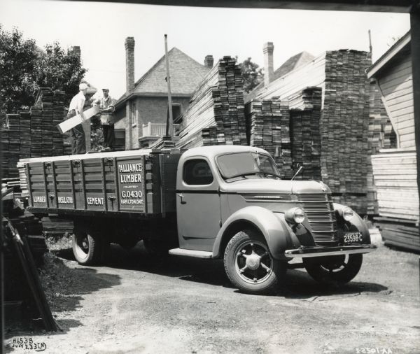 Two men load lumber onto the bed of an International D-35 truck owned by the Alliance Lumber Company Limited.